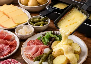 Raclette grill with melted cheese being poured over potatoes, with ham, bacon and grilled vegetables
