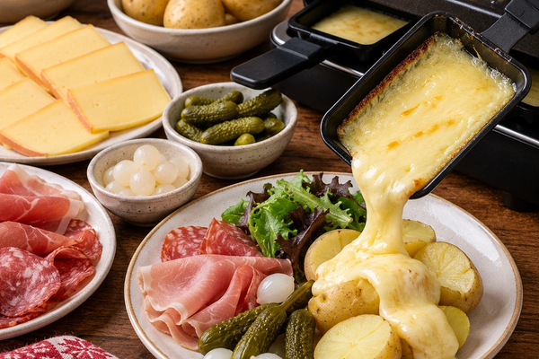 Raclette grill with melted cheese being poured over potatoes, with ham, bacon and grilled vegetables
